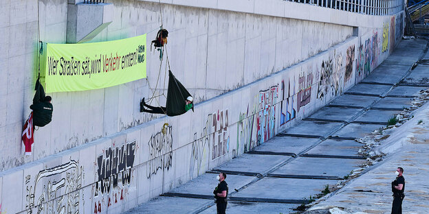 Aktivisten hängen mit einem großen Protestbanner an der Wand über Autobahntrasse. Polizisten stehen unten und schauen nach oben Aktivisten hängen mit einem großen Protestbanner an der Wand über Autobahntrasse. Polizisten stehen unten und schauen nach oben