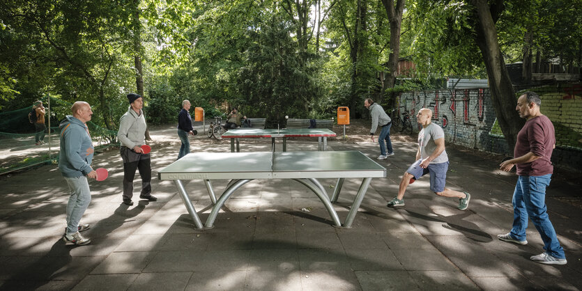 Sechs Männer spielen an zwei Tischtennisplatten, dahinter Bäume