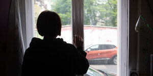 Junger Mensch steht in einem Zimmerfenster und guckt raus