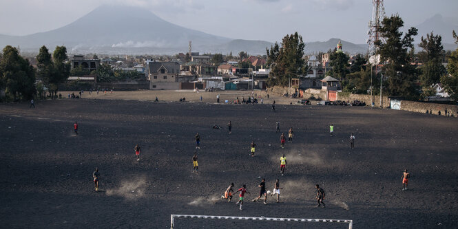 Junge Männer spielen auf einem dunklen Sandboden Fußball vor einem Vulkan