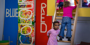 Kindergarten-Kinder stehen auf einer Spielleiter, neben ihnen ist ein Schriftszug auf englisch zu sehen: blue und red. Es is eine bilinguale Kita