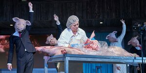 Eva Mattes im weißen Kittel und Haube steht an einem Tisch und säbelt an einem Tierkörper, andere Schauspieler mit Schweinsmasken stehen drumherum