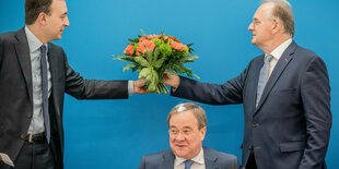 Ein Mann im Anzug übergibt einem anderen Mann im Anzug blumen, dazwischen sitzt ein kleiner Mann im Anzug