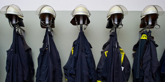 Mehrere Feuerwehrjacken und -helme hängen an einer Garderobe.