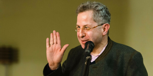 Hans-Georg Maaßen steht hinter einem Mikrofon, lächelt und hebt die rechte Hand