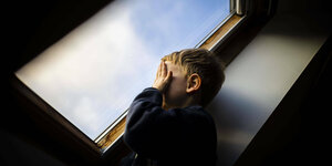 Ein Kind stehtn in einem düsteren Zimmer vor einem Fenster und hält sich eine Hand vor dem Gesicht