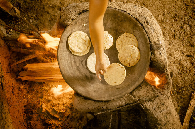 Maistortillas liegen auf einer Platte, die auf einem Feuer steht. Eine Hand dreht die Tortillas um. Es sieht gemütlich und lecker aus