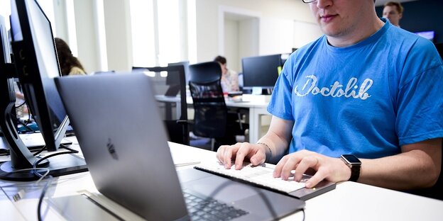 Ein Mann sitzt an einem Laptop und trägt ein blaues T-Shirt mit dem Logo von Doctolib