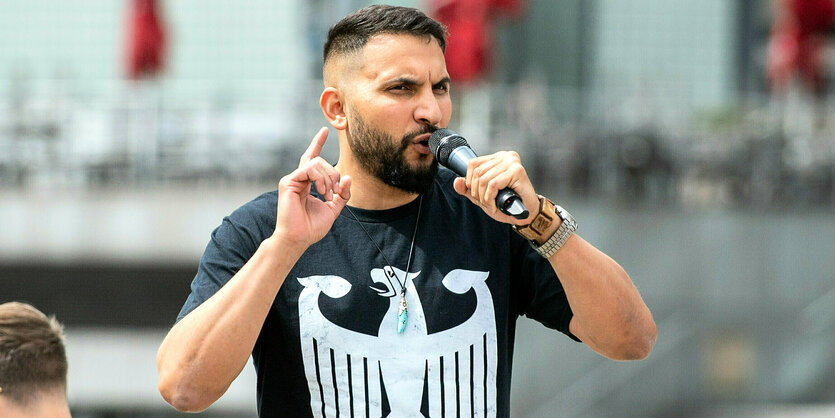 Attila Hildmann spricht in ein Mikrofon und trägt ein schwarzes T-shirt mit weiß aufgedruckten Bundesadler