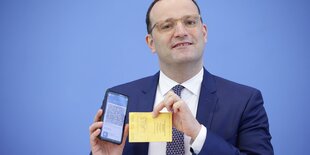 Jens Spahn hält ein Smartphone und einen Impfpass in den Händen