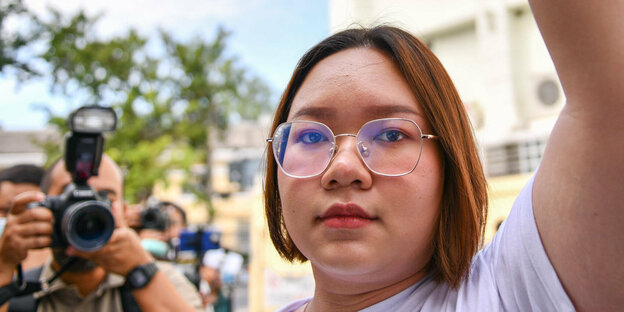 Eine junge Frau mit schulterlangem, rötlichem Haar und Brille, den linken Arm kämperisch erhoben