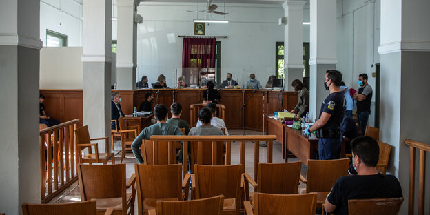 Vier Männer sitzen in der Mitte eines Gerichtssaals, von hinten fotografiert. Auf den Zuschauerrängen sitzt nur eine einzige Person.