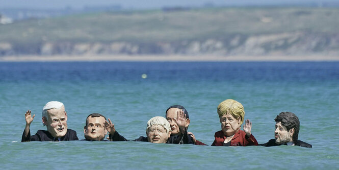 Menschen, die große Masken von Regierungschefs und -chefinnen tragen, sind im Meer. Man sieht nur die Köpfe und winkende arme. Dahinter ein hügeliger Strand.