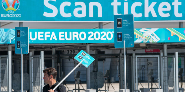 Am Stadion in München hängt ein EM-Werbebanner. Davor steht ein Mann mit Schild