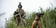 Ein Kommandosoldat des Kommando Spezialkräfte (KSK) der Bundeswehr, steht während eines Videodrehs zum Tag der Bundeswehr mit einem Zugriffsdiensthund, der einen Augen- und Ohrenschutz trägt, in einer Wiese.