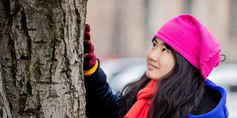 Eine junge Frau mit schwarzen langen Haaren und einer pinken Mütze berührt mit der Hand die Rinde eines Baumes