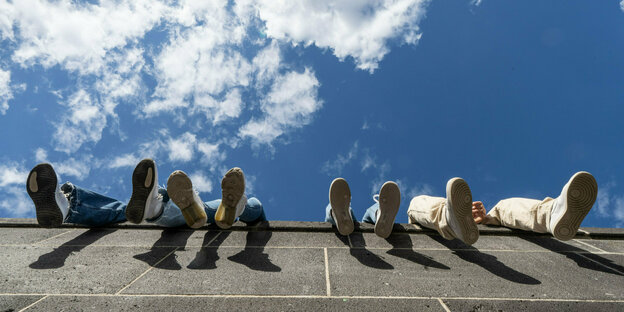 Vier Jugendliche lassen auf einer hohen Mauer vor blauem Himmel mit wenigen Wolken Vier Jugendliche lassen auf einer hohen Mauer vor blauem Himmel mit wenigen Wolken