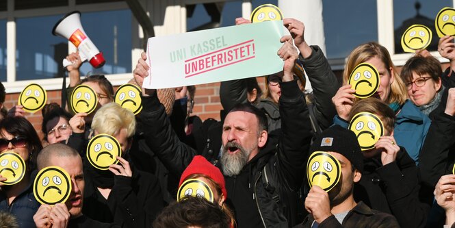 Beschäftigte protestieren vor der Zentralmensa mit traurigen Smileys vor dem Gesicht gegen befristete Verträge an Unis