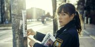 Das Bild zeigt die Schauspieler Verena Altenberger in ihrer rolle als Polizeioberkamissarin Elisabeth Eychhoff im Polizeiruf aus München. Sie klebt ein Vermissenblatt an eine Laterne