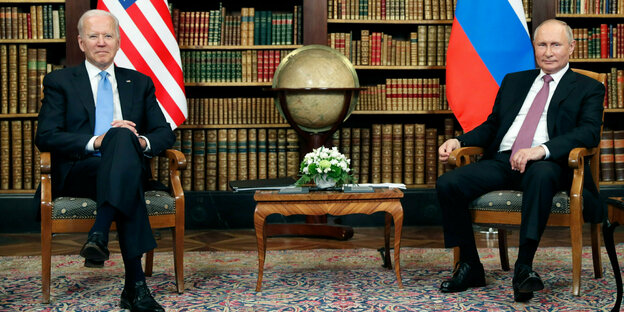 Der russische Präsident Wladimir Putin und US-Präsident Joe Biden sitzen bei ihrem Treffen vor Bücherregal, Landesflaggen ihrer Länder und einem Globus