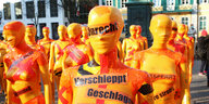 In orangefarbene Folie eingewickelte Gestalten mit Klebestreifen mit der Aufschrift "bedroht, verfolgt"