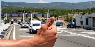 Auf dem Foto sieht man einen Daumen, der nach oben gestreckt ist, im Hintergrund eine mehrspurige Straße mit Autos