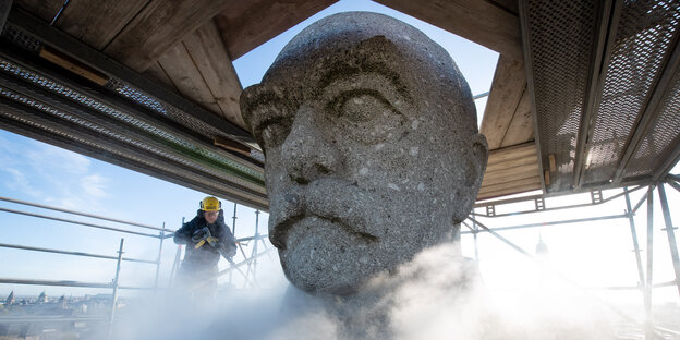 Ein Arbeiter reinigt einen überlebensgroßen Kopf mit einem Dampfstrahler