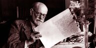 Sigmund Freud sitzt an seinem Schreibtsich und liest ein Manuskript