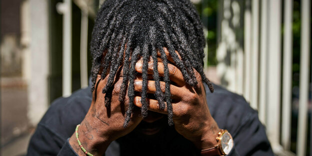 Ein Mann mit schwarzer Hautfarbe hält sich die Hände vors Gesicht, seine Haare fallen über die Hände