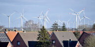 Das Bild zeigt Windräder hinter einem Wohngebiet, in dem manche Häuser Solaranlagen auf dem Dach haben.