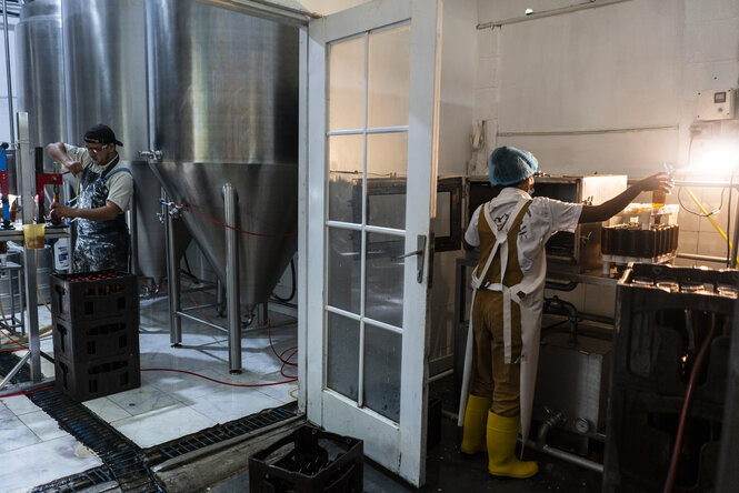 Die Brauerei von innen. Es ist eine ehemalige Kirche. Links steht ein Mann und setzt mit einer Maschine Kronkorken auf die Flasche. Rechts führt eine offene Tür in einen anderen Raum. Dort steht eine Frau und stabelt gereinigte Flaschen in einen Kasten.