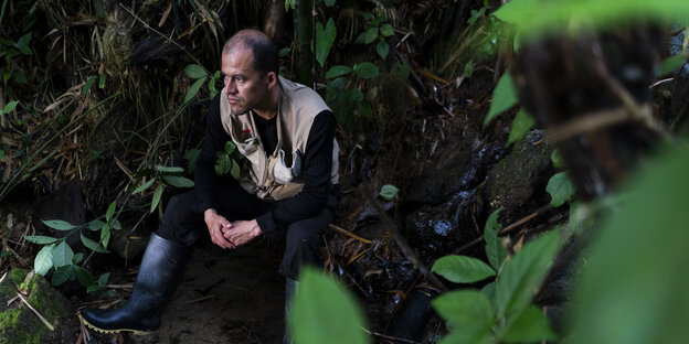 Lorenzo Bautista sitzt neben einer kleinen Quelle in Minca, Kolumbien. Er ist ein Mann mittleren Alters mit lichtem, kurzen Haar. Er trägt Regenstiefel, einen schwarzen Pullover und eine Weste mit vielen Taschen. Bautista blickt grimmig, aber entschlossen