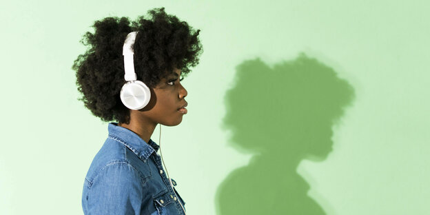 Eine junge Frau trägt Kopfhörer. Dahinter ist eine grüne Wand, auf der man ihren Schatten sieht.