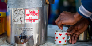 Ein Mensch rührt in einer Tasse neben einem Warmhaltebehälter für Tee oder Kaffee. Darauf steht "kein Mensch ist illegal"