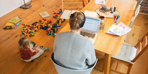 Ein zweijähriges Kind spielt im Wohnzimmer, während seine Mutter Zuhause im Homeoffice an einem Laptop arbeitet.