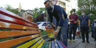 Alessandro Zan streicht eine Bank im Freien in Regenbogenfarben. Zan ist ein Mann mittleren Alters mit kurzen dunklen Haaren und Bart. Er trägt eine Maske