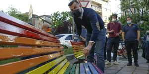 Alessandro Zan streicht eine Bank im Freien in Regenbogenfarben. Zan ist ein Mann mittleren Alters mit kurzen dunklen Haaren und Bart. Er trägt eine Maske