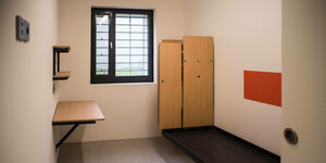 Eine Gefängniszelle von Innen. Darin stehen eine Pritsche und ein Schrank. An der Wand sind ein kleiner Tisch und zwei Regalbretter befestigt. Das Fenster ist vergittert.