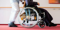 Ein Pfleger schiebt eine Bewohnerin eines Pflegeheims in einem Rollstuhl über den Flur.