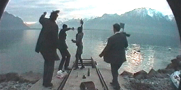 Eine Gruppe Musiker:innen steht mit dem Rücken zur Kamera auf einer Brücke an einem See und scheint zu feiern