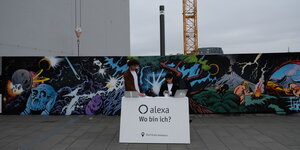 Zwei junge Menschen sitzen an einem Tisch vor einem Bauzaun mit Graffiti: Auf einem Schild steht: "Gratis Audiotour"