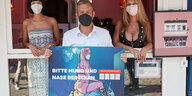 Ein Mann mit schawrzer Maske hält eine Tafel mit dem Slogan "Bitte Mund und Nase decken" und einer gezeichneten Frau, die auf dem Gesicht eines Mannes sitzt. Im HIntergrund stehen zwei Frauen an Fenstern