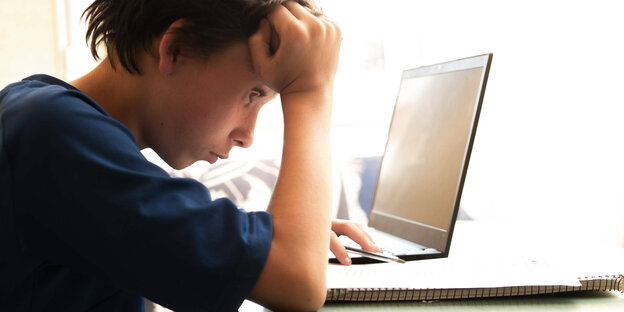Ein Junge sitzt zuhause vor Laptop und einem Schreibblock - den Kopf mit der Hand abgestützt