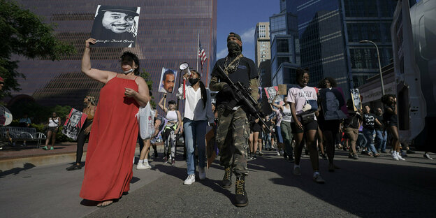 Eine Gruppe von Demonstrierenden in Minneapolis. Sie tragen ein Schild über sich mit einem Opfer von Polizeigewalt darauf. Ein bewaffneter Security ist dabei.