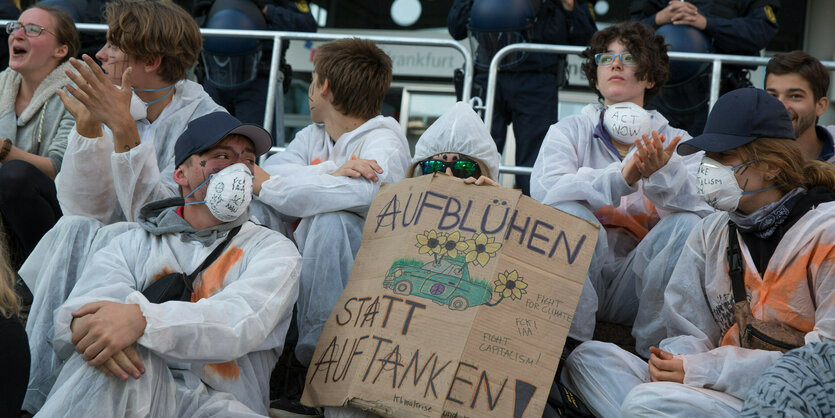 Demonstranten in weißen Overals sitzen vor einer Absperrung. Auf einem großen Karton steht "Aufblühen statt Auftanken"