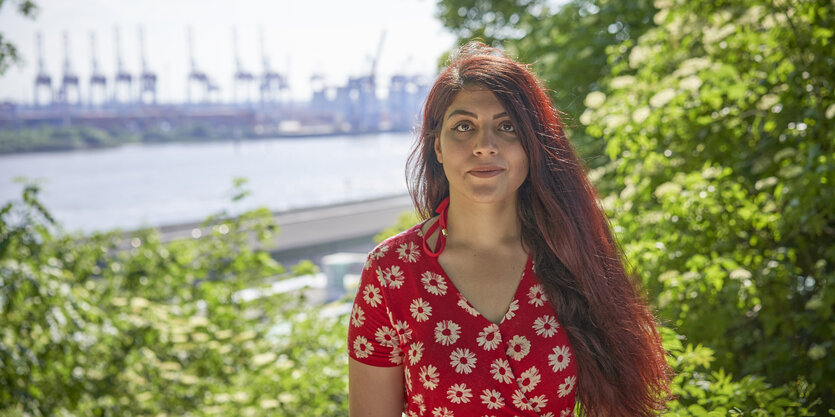 Eine Frau im roten Kleid steht vor Bäumen, dahinter die Elbe und Kräne