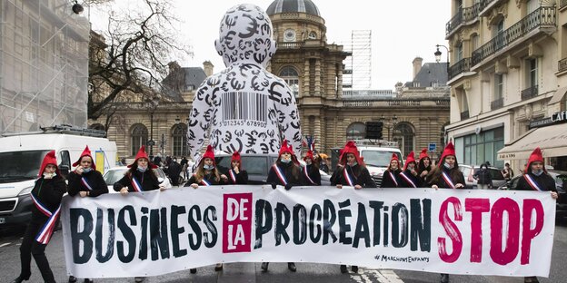 Frauen mit roten Mützen tragen ein Banner mit der Aufschrift "Business de la Procration STOP" vor einer großen aufgepumpten Babyfigur mit aufgedrucktenEurozeichen