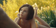 Elli (Lena Watson) liegt im Gras und sieht mit wächsernem Gesicht hoch, vor ihr ragt ein Männerarm ins Bild.