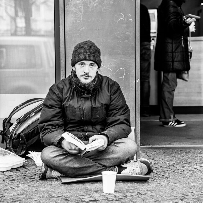 Junger Mann mit Mütze sitzt mit gekreuzten Beinen auf der Straße, vor sich ein Plastikbecher für Spendengeld