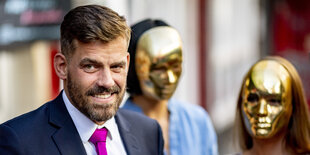 Ein Mann mit pinker Krawattem im Hintergrund zwei Frauen mit goldenen Masken im Gesicht